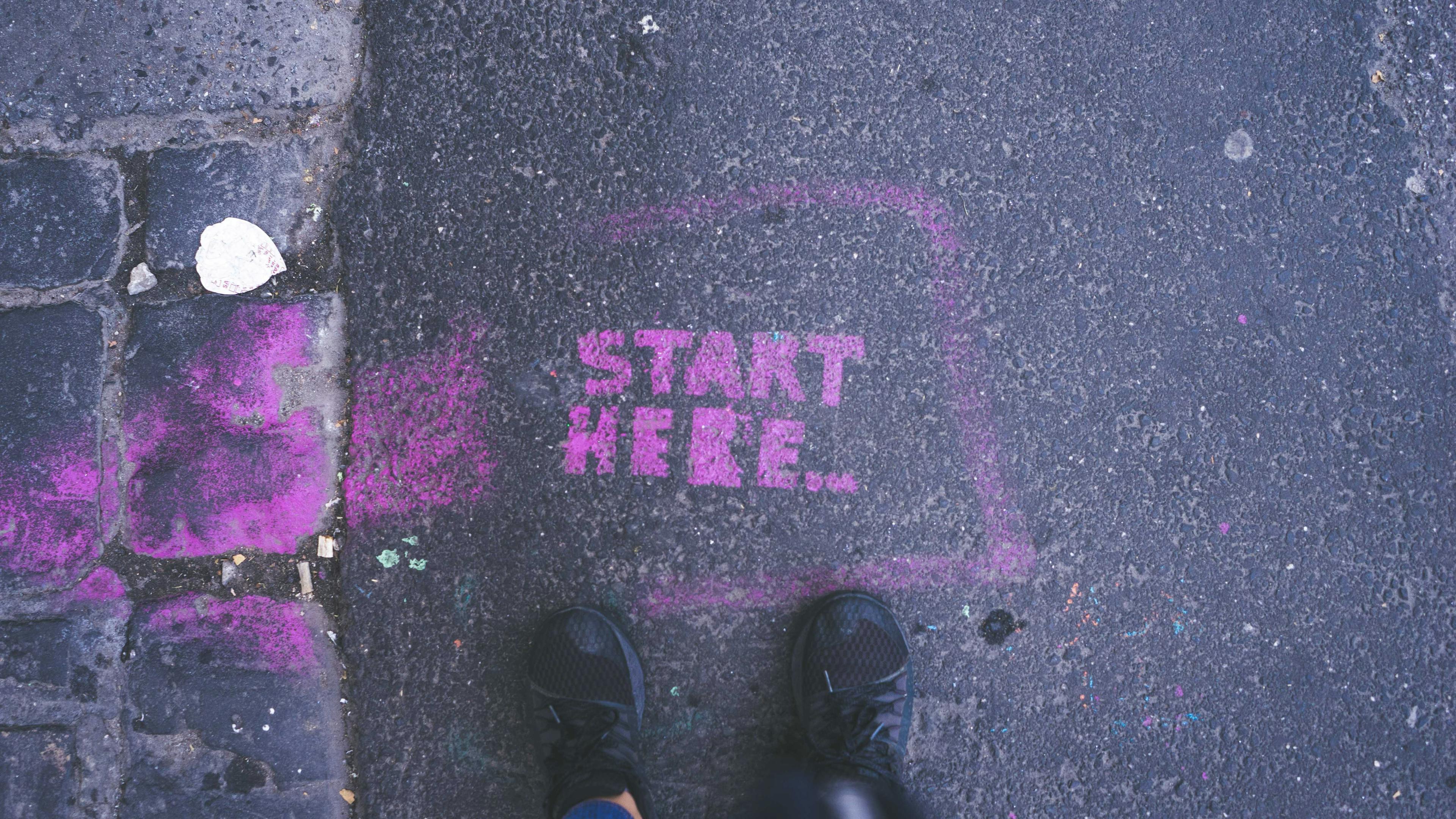 The words "start here" in purple spray paint on a street with feet near it.