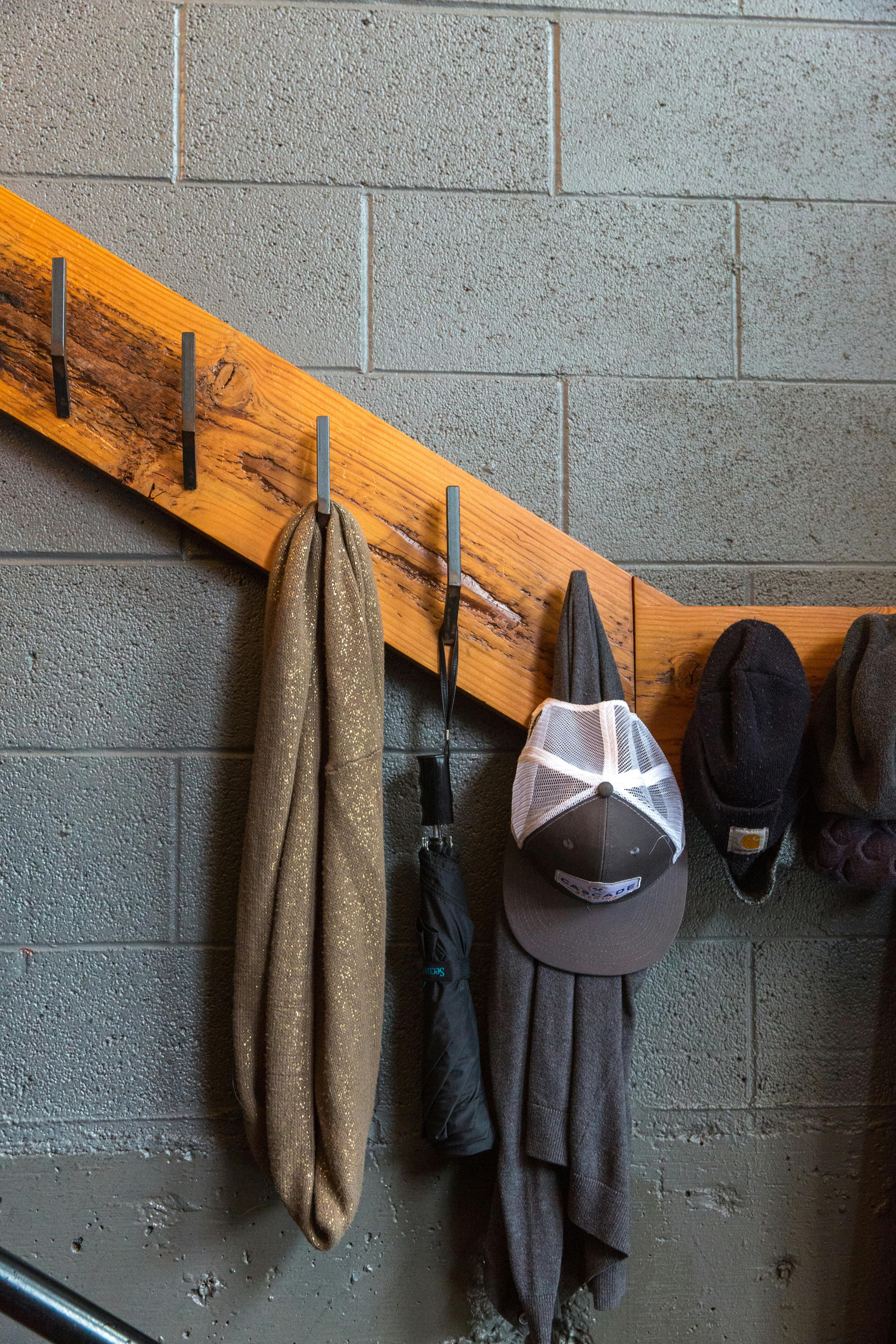 A scarf, umbrella, and hat hanging from metal hooks attached to a wooden rail on a cement block wall